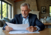 Taux d’imposition sur la vente d’une résidence secondaire : lois et conseils à connaître Homme d'âge moyen lisant des documents immobiliers dans un salon lumineux